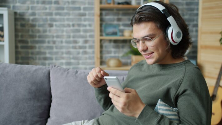 Young man wearing headphones uses smartphone on couch.
